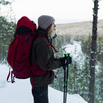 En el norte de Finlandia, bajo la suave luz invernal, una excursionista con mochila roja y bastones de trekking contempla desde una cumbre nevada un valle cubierto de bosque.