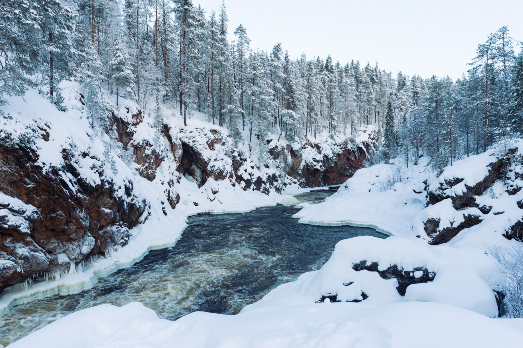 A partially frozen river winds between steep, snow-laden banks and frost-covered trees in the Kiutaköngäs rapids in northern Finland.