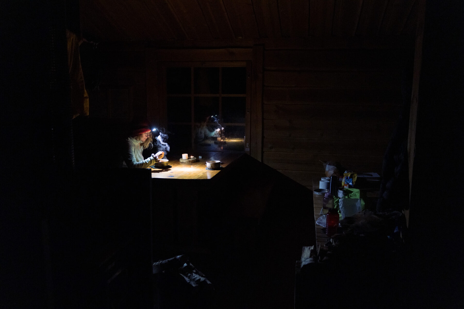Headlamp beams light a small table as two hikers eat inside a dim wooden shelter at night.