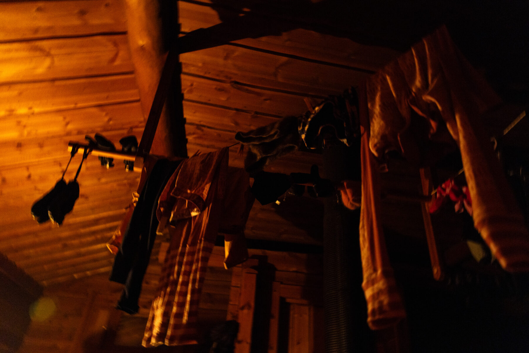 Clothes and gloves hang from wooden beams inside a warmly lit cabin, drying by the heat.