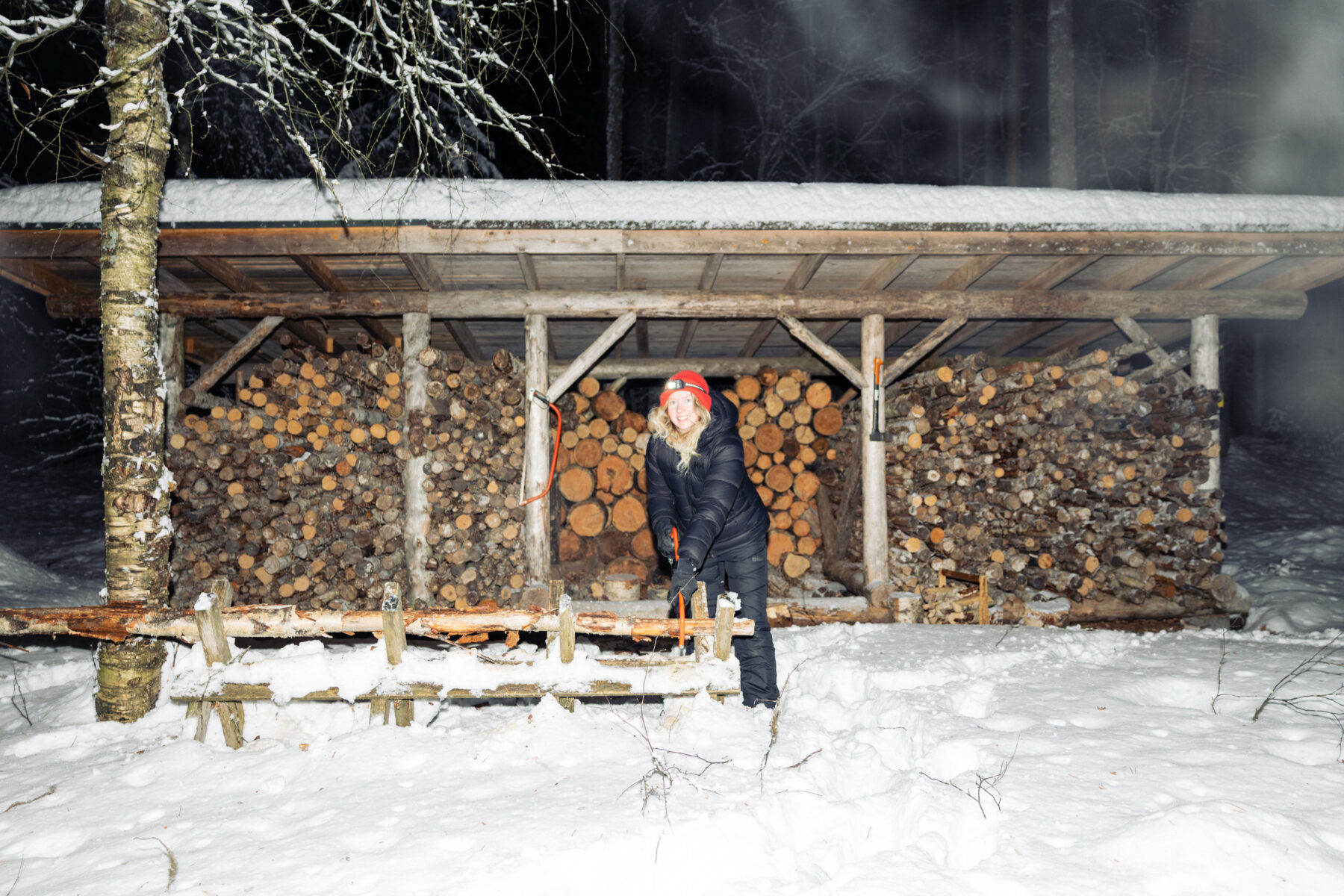 A hiker in winter gear prepares firewood outside a wooden shelter surrounded by snow and stacked logs, in northern Finland.