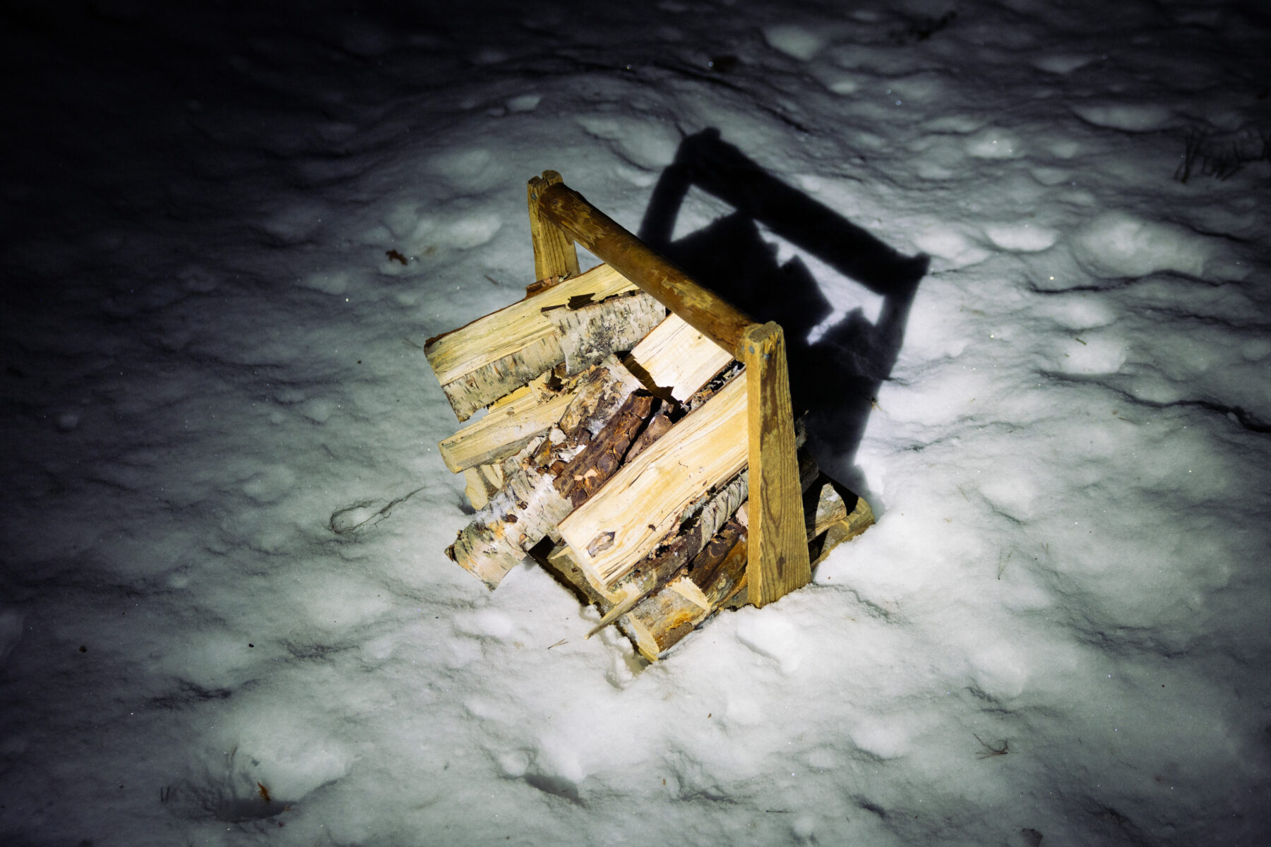 A small wooden basket filled with chopped firewood stands on snow at night, lit by a headlamp that casts a sharp shadow.