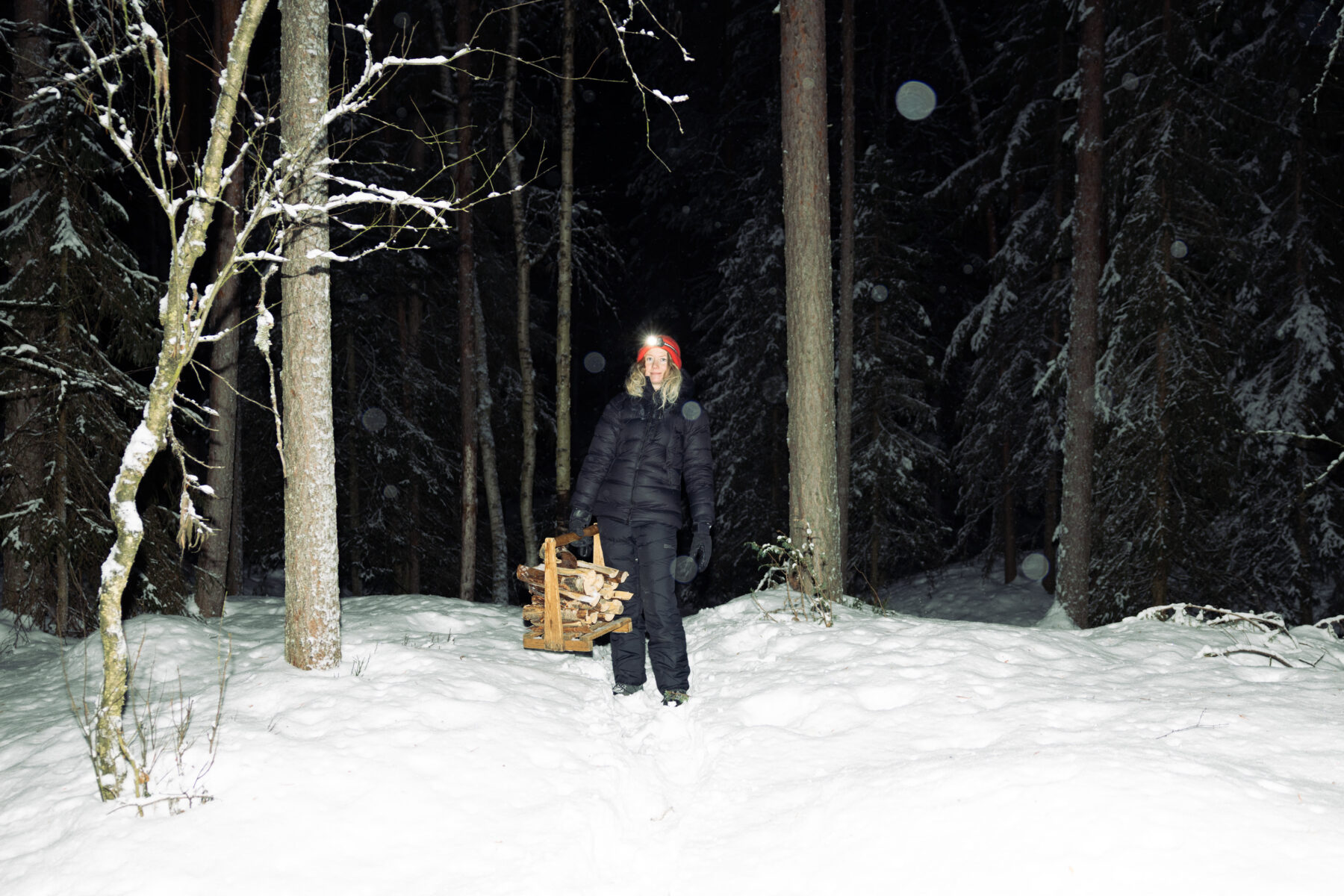 A hiker wearing a red hat carries firewood across a snowy clearing at night, surrounded by tall trees.