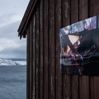 A photographic artwork by Lada Suomenrinne hangs on the dark wooden wall of a coastal building, with snowy hills and grey sea stretching into the distance.