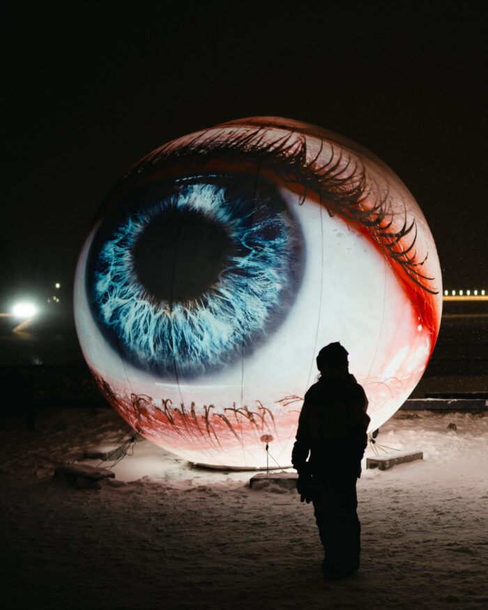 A person stands before a giant illuminated sculpture of a blue human eye displayed outdoors at a light art festival.