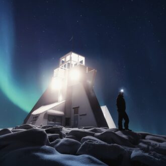 A lone figure wearing a headlamp stands beside a lighthouse as aurora lights ripple through the dark sky.