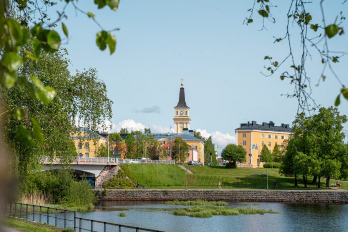 A riverside view of Oulu shows historic buildings and a church tower rising above green parkland on a clear day.