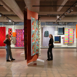 Zwei Besucher betrachten farbenfrohe finnische Textilien, die in einem weitläufigen Raum der EMMA-Ausstellung „Draped“ aufgehängt sind.