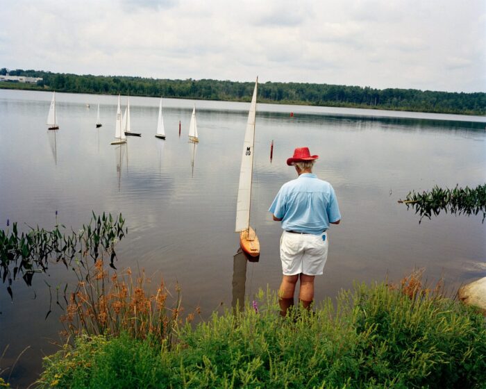 A man in a red hat guides a radio-controlled sailboat on a calm lake with other small boats nearby.