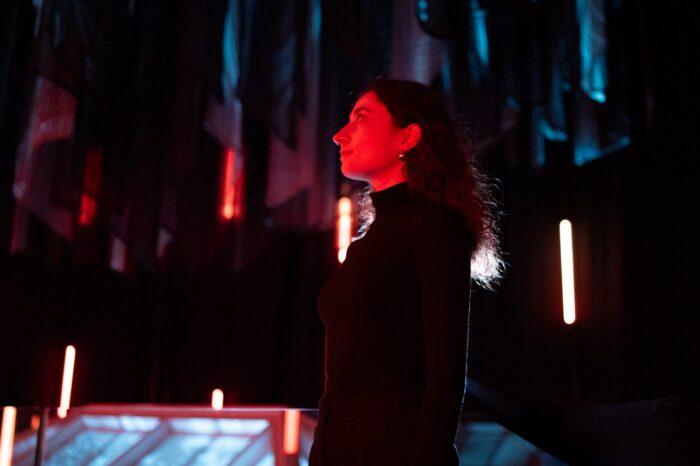A woman gazes upward in a darkened space lit by red and blue lights, surrounded by suspended materials and vertical light strips.