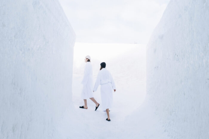 Two people in bathrobes and slippers walk down a hallway whose walls, floor and ceiling are made of snow and ice.