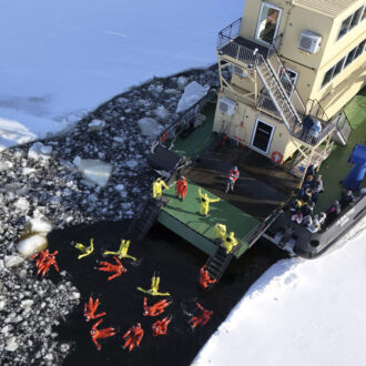 In a view from above, a large ship floats in a channel between sheets of ice, while people in red protective suits float on their backs in the water.