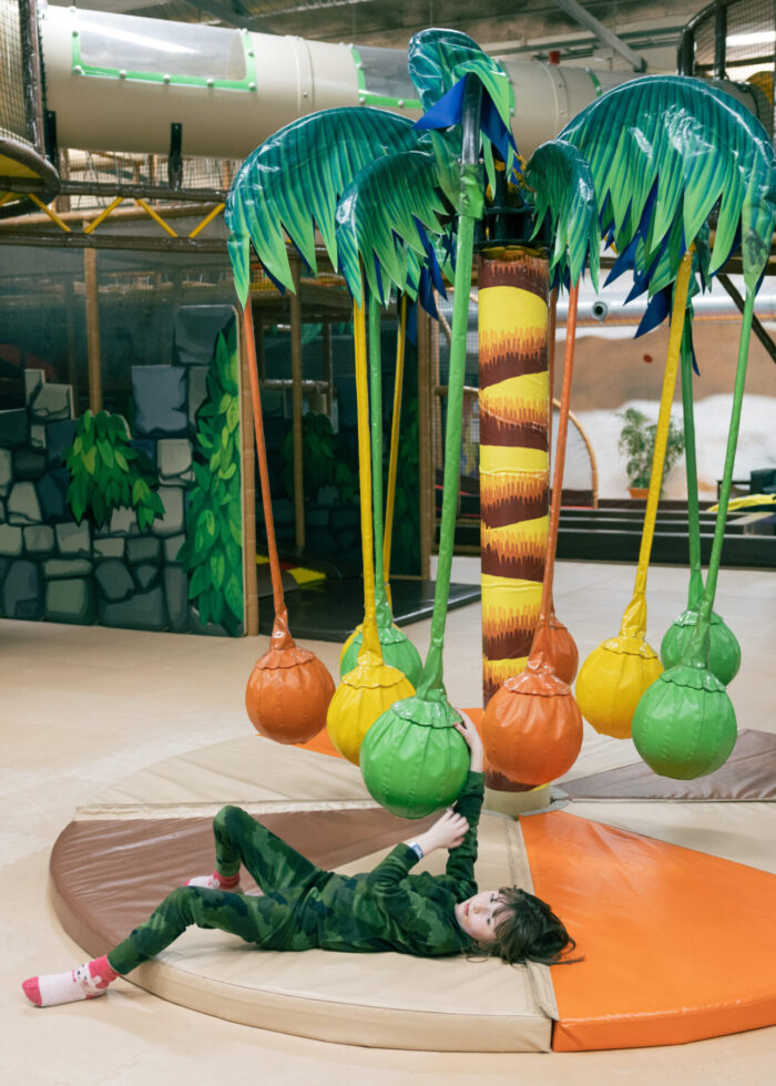 A child plays on a padded floor under a palm tree-style soft play structure at an indoor playground.