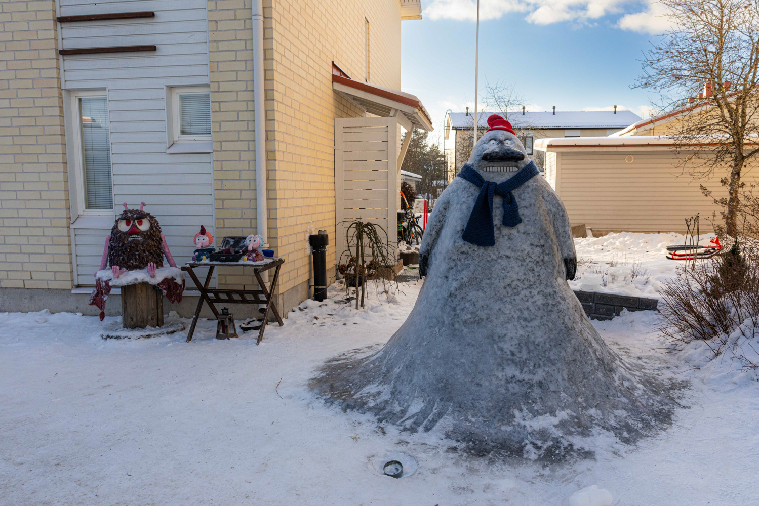 A large snow sculpture of the Groke, a Tove Jansson Moomin character who has a stern face and whose feet are hidden by a long gown, stands in the yard in front of a house.