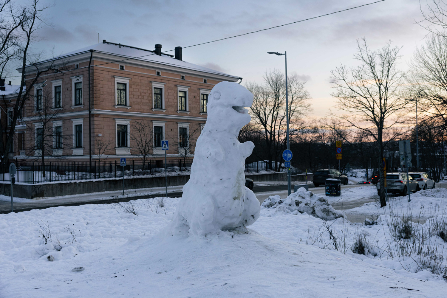 A snow sculpture of a dinosaur stands on a small hill beside a road and a brick building.
