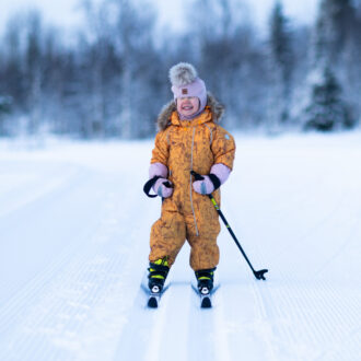 A young child in a yellow snowsuit and pink hat smiles while standing on skis on a groomed ski track with snowy trees in the background.