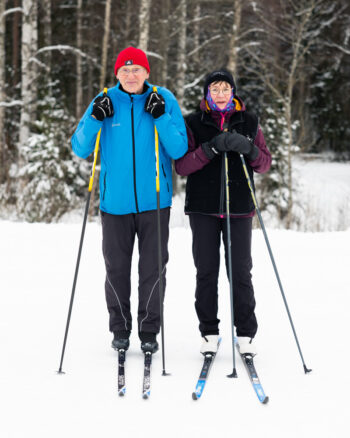 An older man and woman in winter clothing pose on skis in the snow with trees behind them.