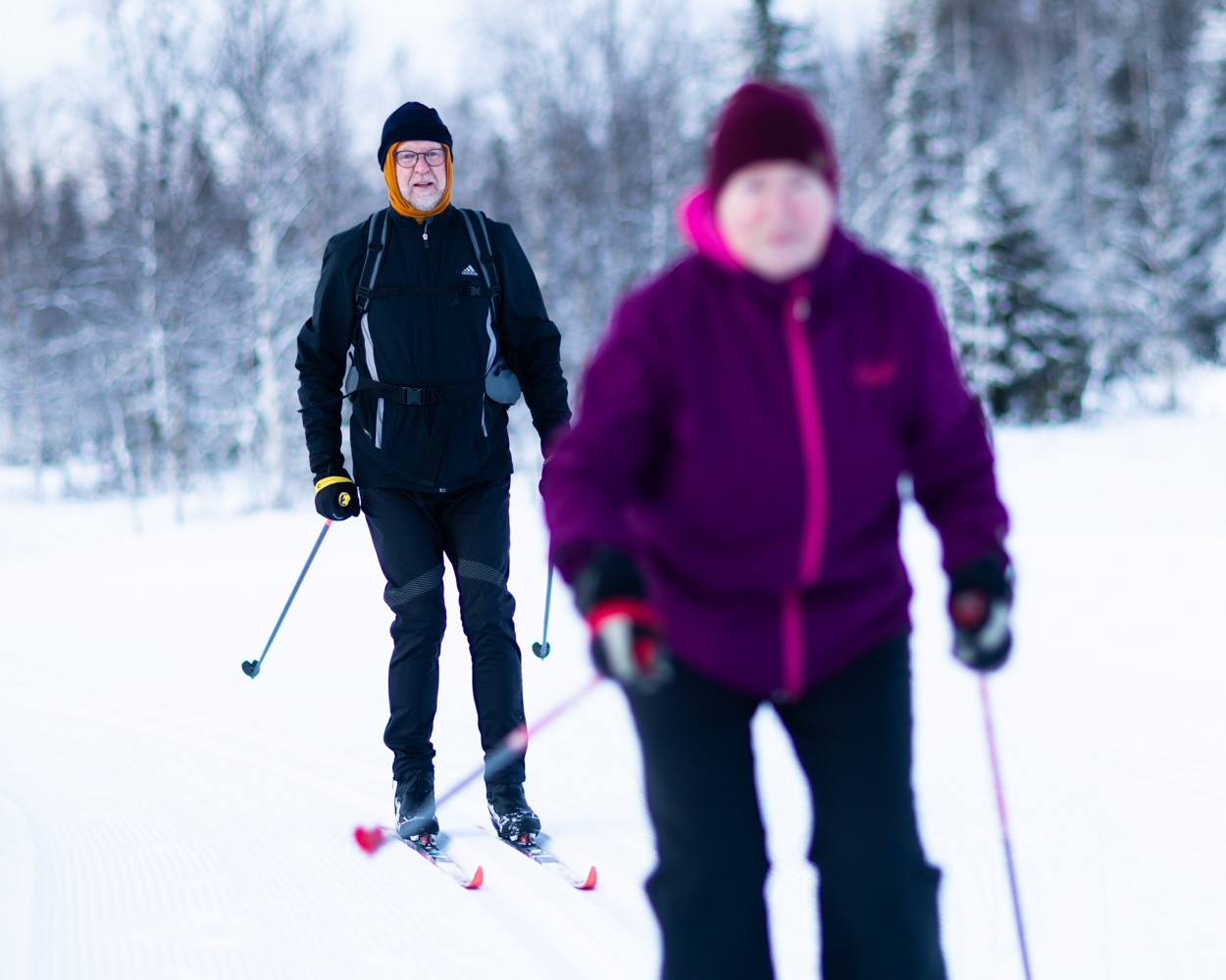 A man skis along a snowy trail behind a woman out of focus in the foreground, both wearing winter clothing.