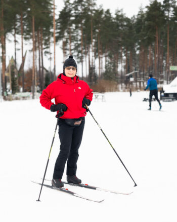 A cross-country skier in winter clothing stands with ski poles, with pine trees and another skier behind.