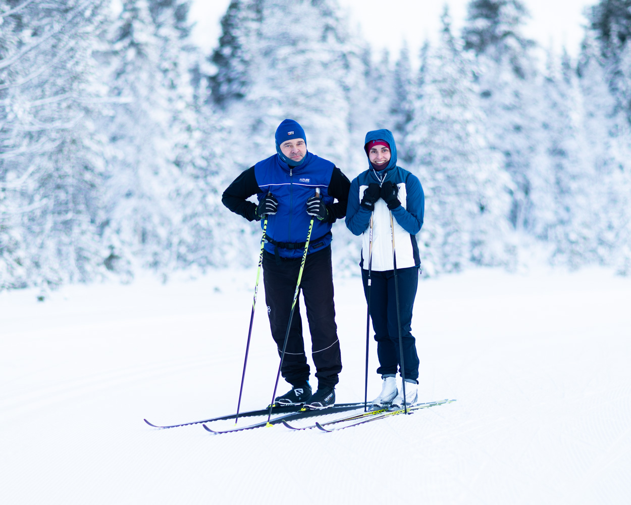 A man and woman stand on cross-country skis with trees heavy with snow behind them.