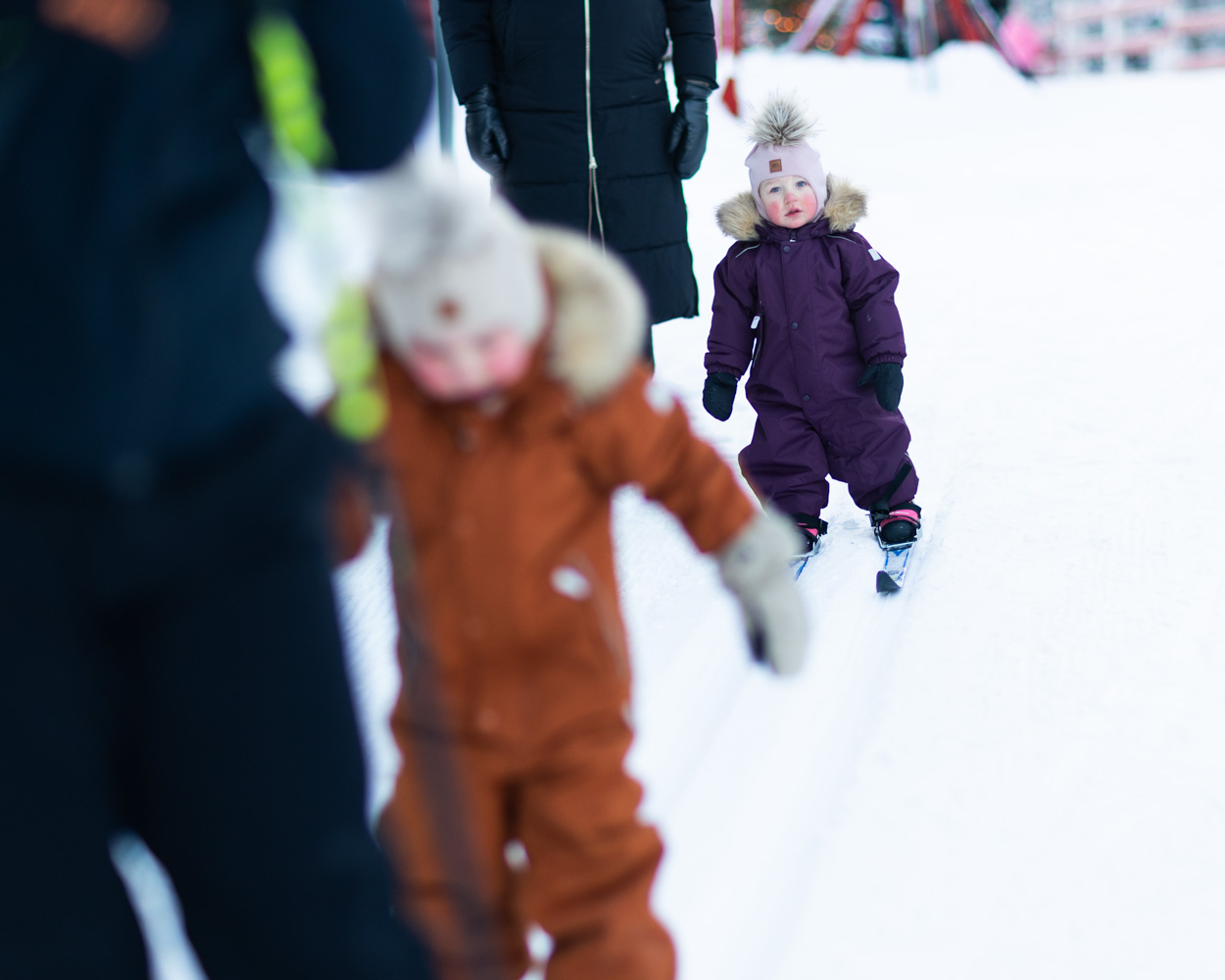 Young children dressed in warm snowsuits and hats ski along a track in the snow with adults nearby.