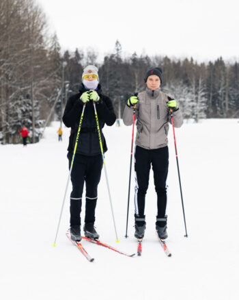 Two people stand on cross-country skis holding poles on a snowy field with trees in the background.