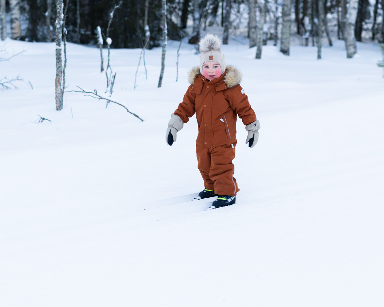 A small child in a brown snowsuit skis across snow with trees in the background.