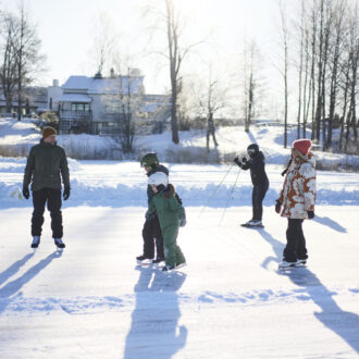 Families skate along a marked track on a frozen lake under bright winter sun, with long shadows stretching across the snow.