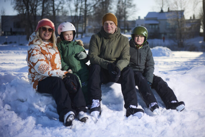 Two adults and two children sit together on a pile of packed snow beside a frozen lake, dressed in winter coats and helmets, with ice skates on their feet.