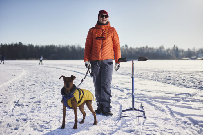 A woman in winter clothes stands on a snowy track beside a dog on a lead and a kicksled, under a clear winter sky.