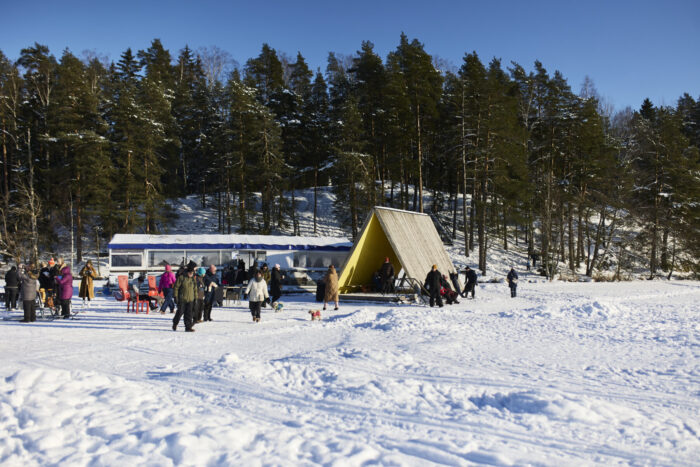 People gather near a small building at Fjällbo Jetty on a snowy lakeshore, with pine trees rising behind the frozen lake.