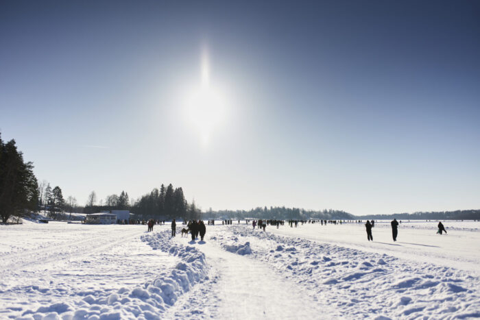 People stroll and skate across a wide frozen lake on a sunny winter weekend, following snowy tracks under a clear sky.