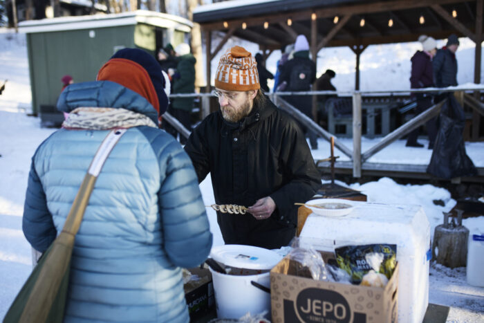 A man in winter clothing serves food to a customer at an outdoor grill on a snowy lakeshore.