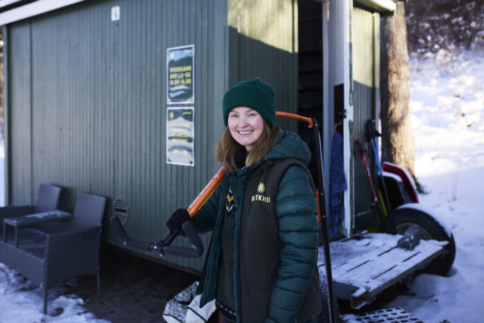 A winter guide smiles while holding a kicksled, a sled that has handlebars, outside a small lakeside hut, with snow on the ground and more gear by the entrance.