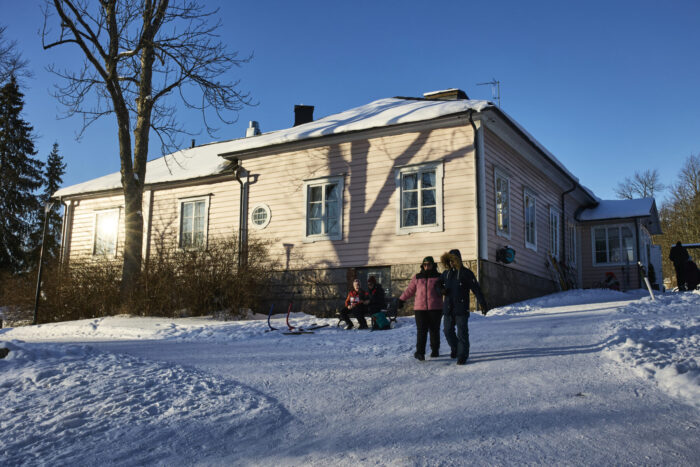 People walk along a snow-covered path beside a wooden café building near a frozen lake, while others sit outside in the sun.