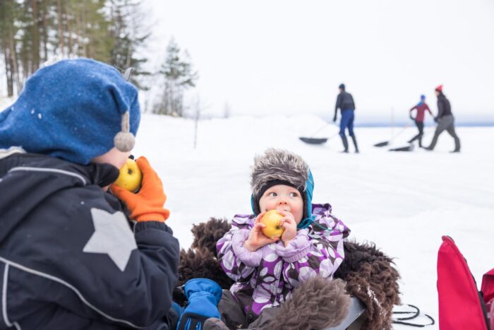 Two small children in winter clothes eat apples while adults shoveling snow are visible in the background.
