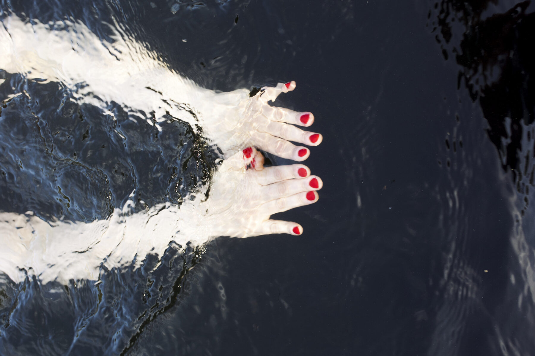 Two hands and arms with bright red nail polish on the fingernails are in water.