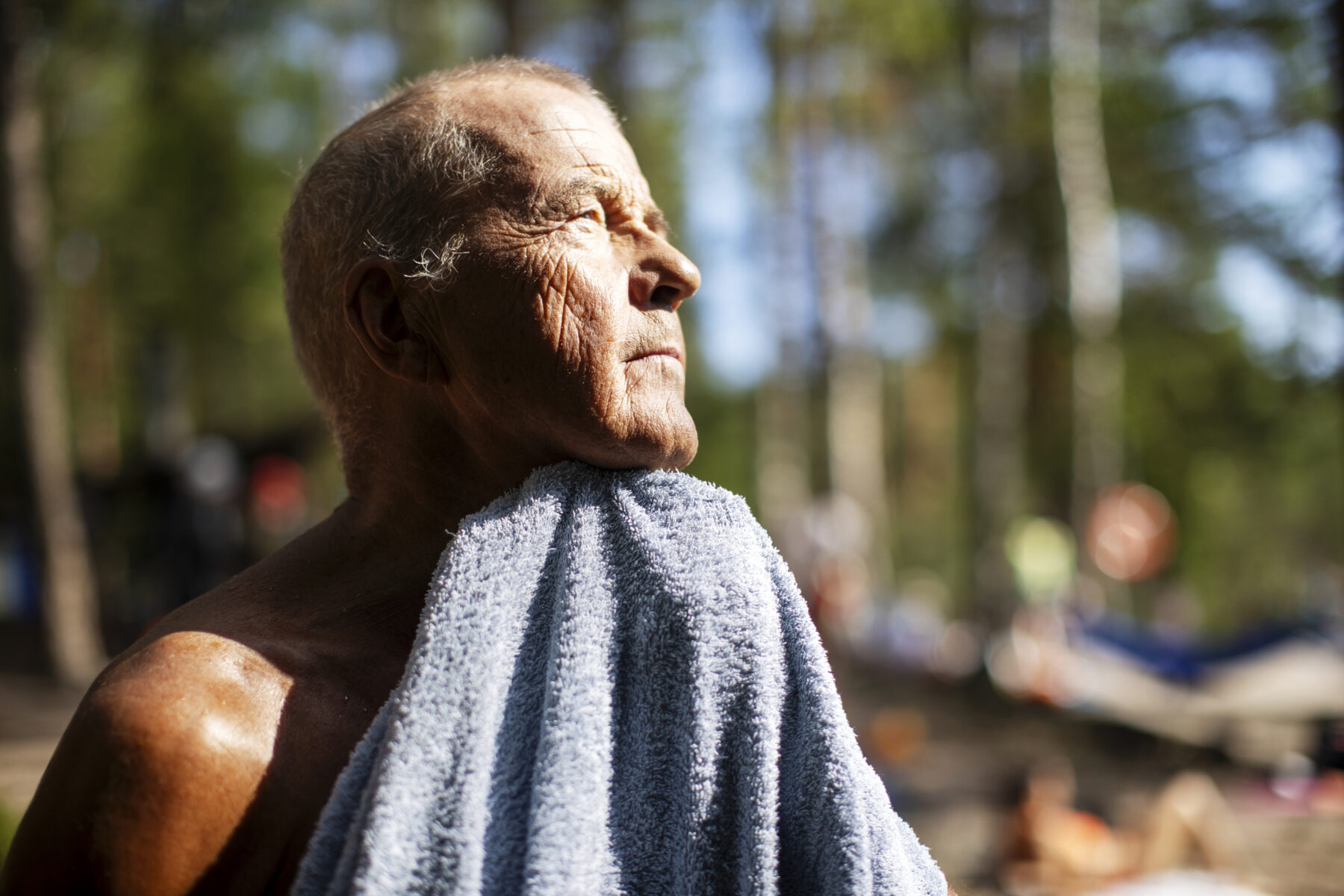 A man, Martti Laajus, looks into the sun as he dries himself with a towel. Trees are in the background.