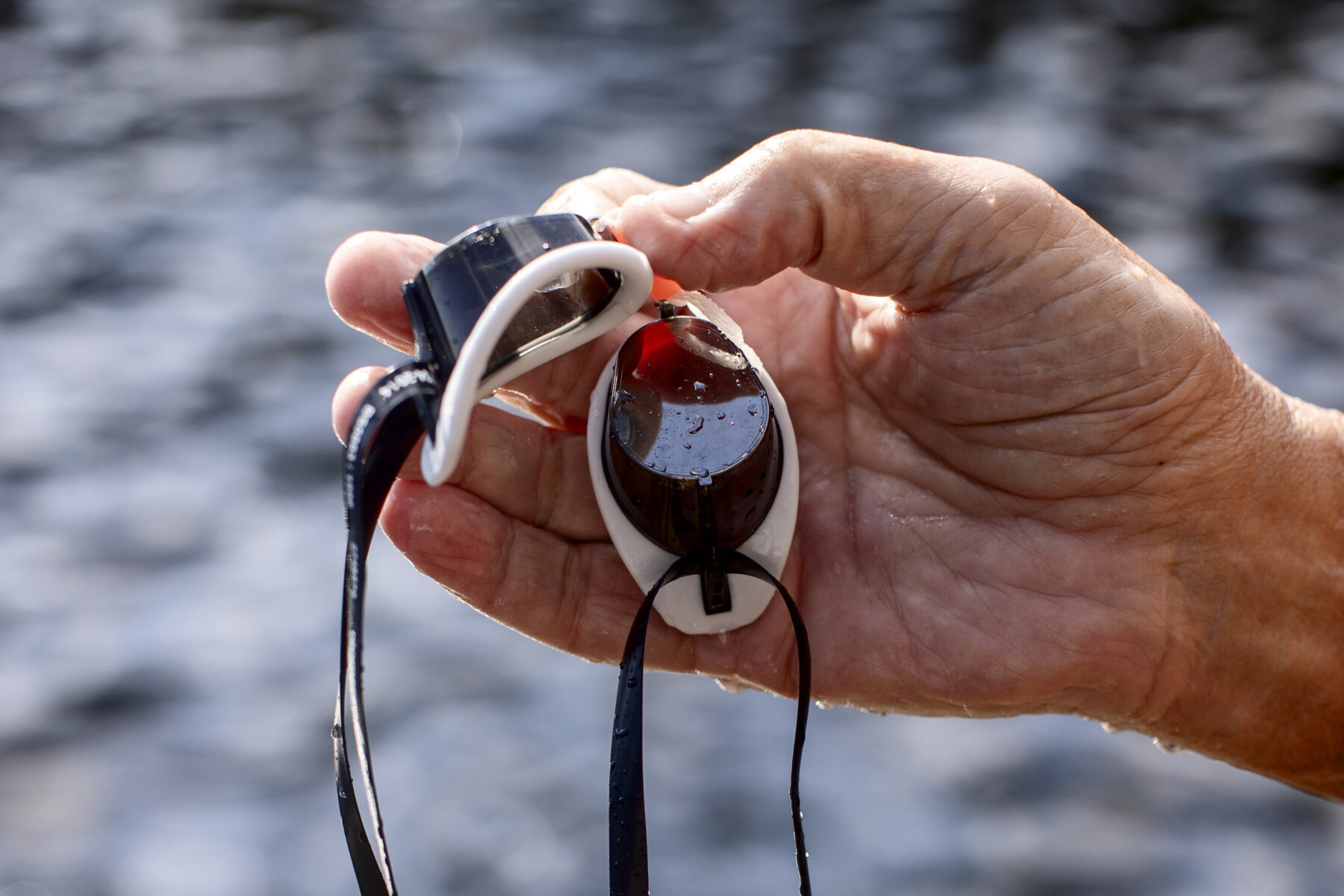 A hand holds swimming goggles with water in the background.