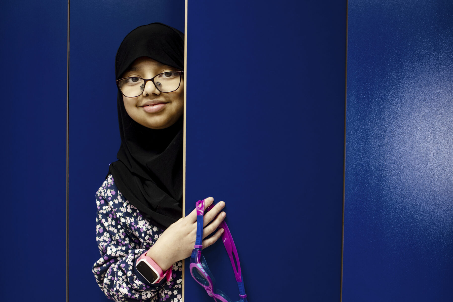 A woman, Aisha Siddiqi, looks around the edge of a door as she holds swimming goggles.
