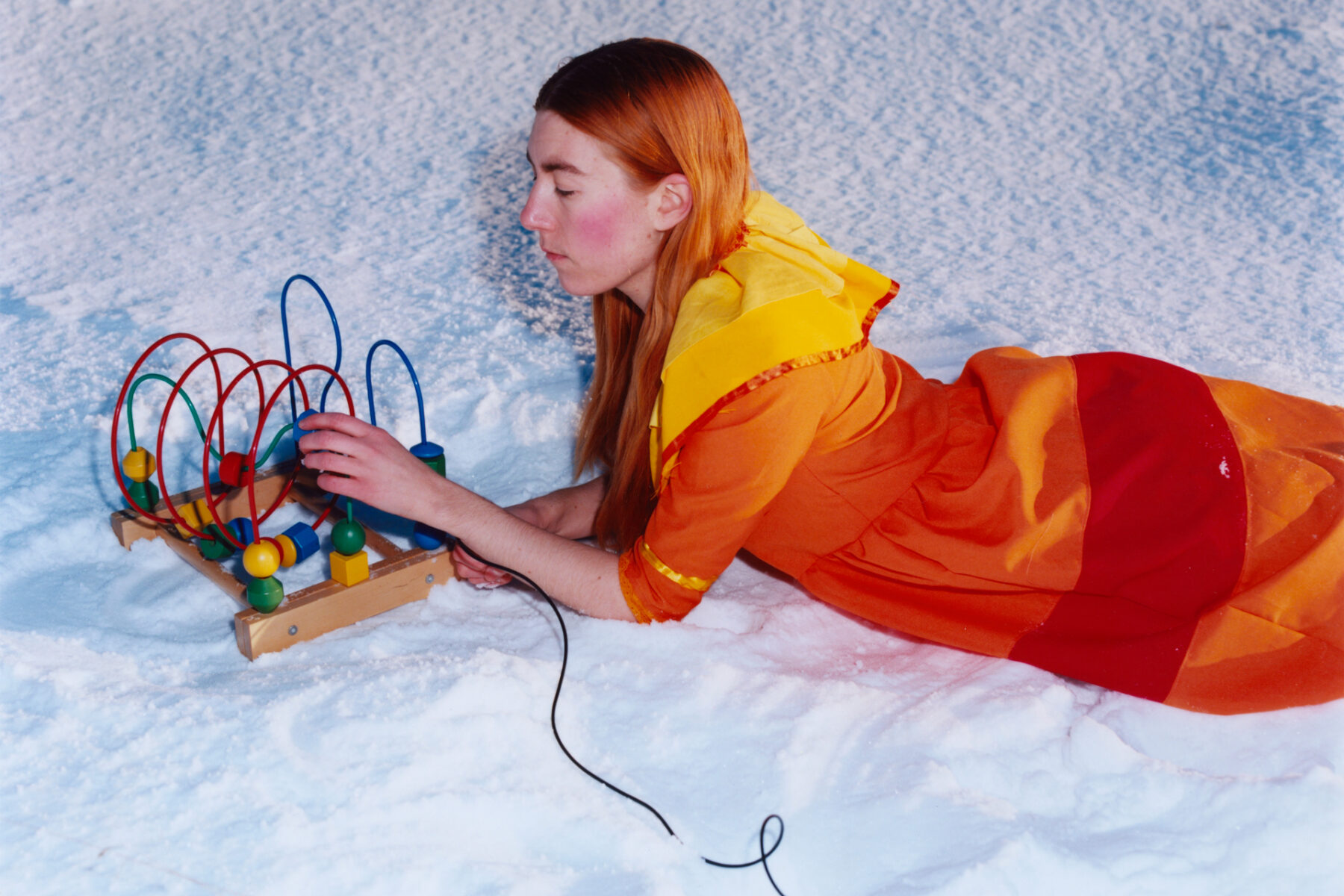 Une femme, en l'occurrence Emma Sarpaniemi, est allongée sur le ventre au milieu de la neige, en train de manipuler un jouet pour bébé. Elle porte une robe jaune, orange et rouge.
