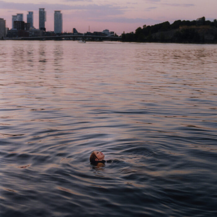 Eine Frau schwimmt in einem Gewässer und blickt nach oben. Hochhäuser und eine Brücke sind im Hintergrund zu sehen.