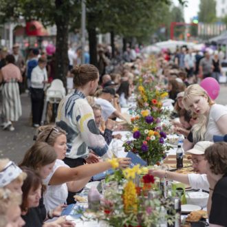 Many people eat at a long table outdoors. The table is filled with plates, food and flowers.