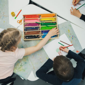 In a view from above, several children are shown sitting at a table drawing pictures of trees with coloured pencils.