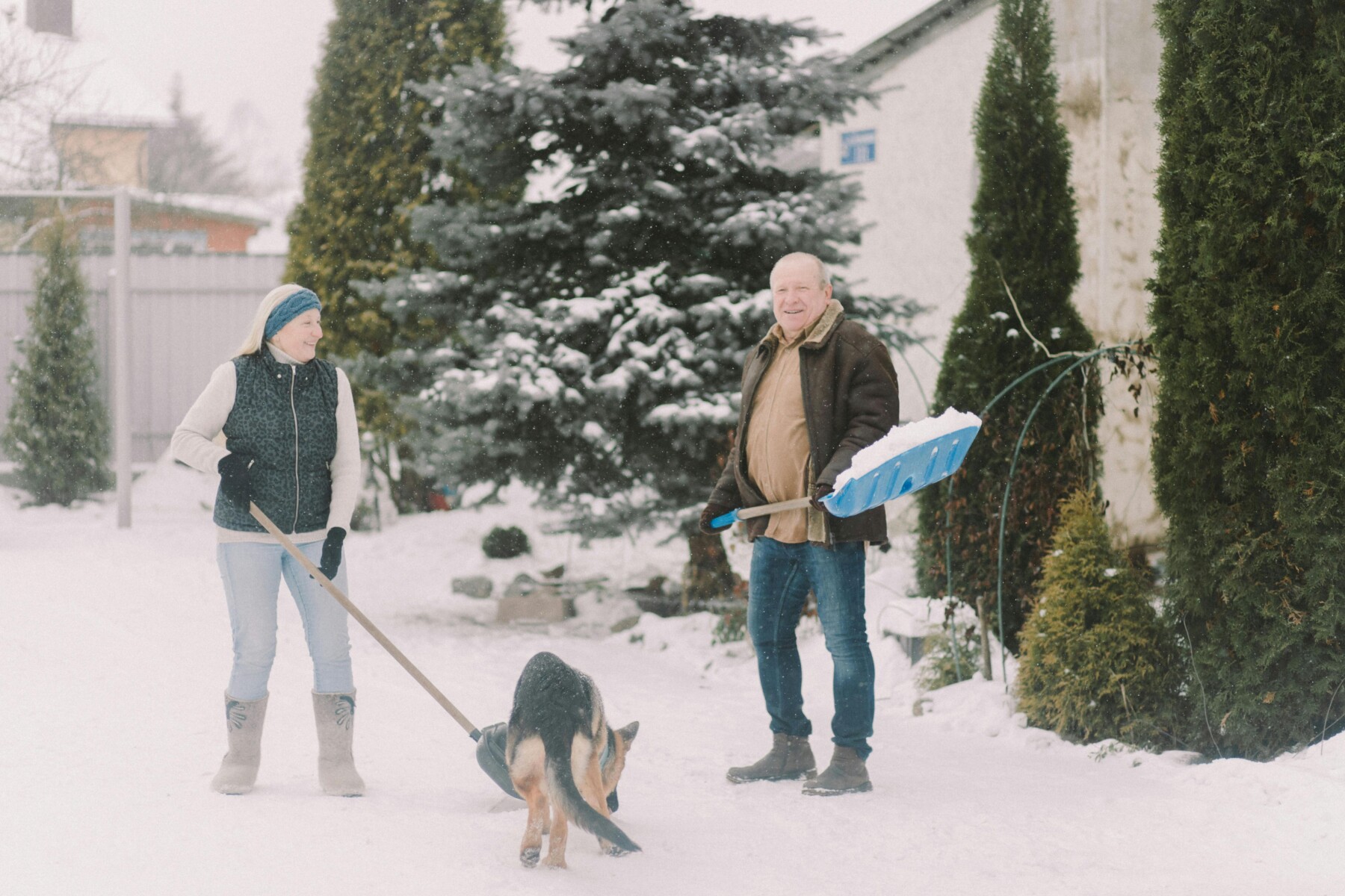 A woman and a man shovel snow side by side in a wintery yard while a dog runs between them.