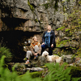 In a nature area in Helsinki, Finland, two women and a dog are standing in front of a wall of rock flanked by trees and moss.