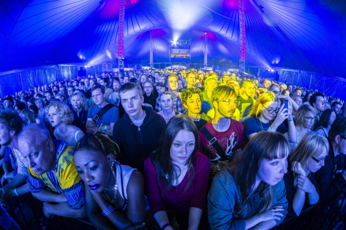 A large crowd of concertgoers stands packed together under blue, yellow and purple stage lighting inside a tented music venue in Finland.