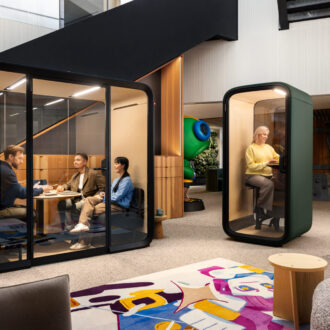 Des employés sont en train d’utiliser les cabines de bureau insonorisées de la société finlandaise Framery dans un open space, avec d’un côté une réunion à trois participants qui se tient dans une grande cabine, tandis que du côté opposé de l’espace, une personne seule travaille dans une cabine de plus petites dimensions.