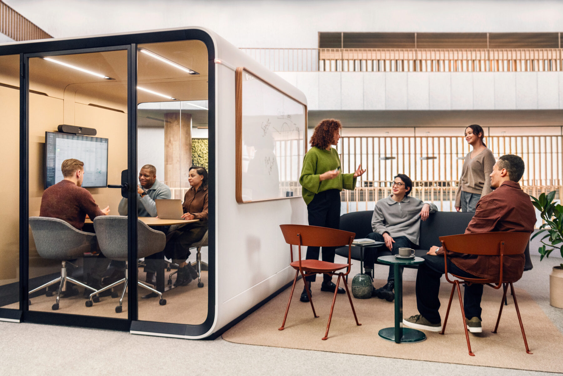 Trois employés discutent à l’intérieur d’une cabine de bureau vitrée conçue par la société finlandaise Framery, tandis qu’à droite de cette cabine, des collègues à eux tiennent une réunion dans un espace de travail ouvert et convivial, certains d’entre eux semblant avoir les yeux tournés vers un tableau blanc fixé sur la paroi extérieure de la cabine.