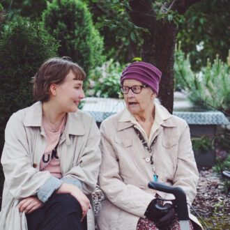 A young woman and an elderly woman sit on a garden bench, smiling and talking, with a walker beside them.